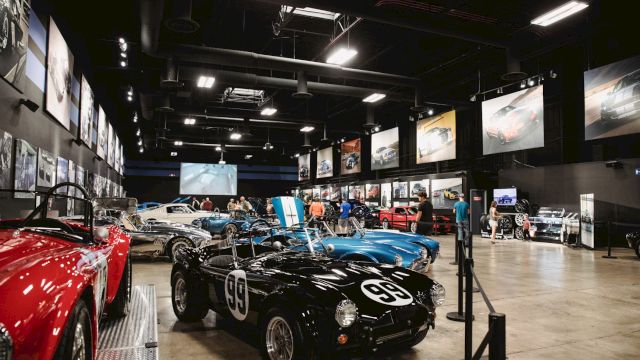 The image shows a car museum exhibit with classic cars, including a black one labeled "99," surrounded by visitors and automotive displays.