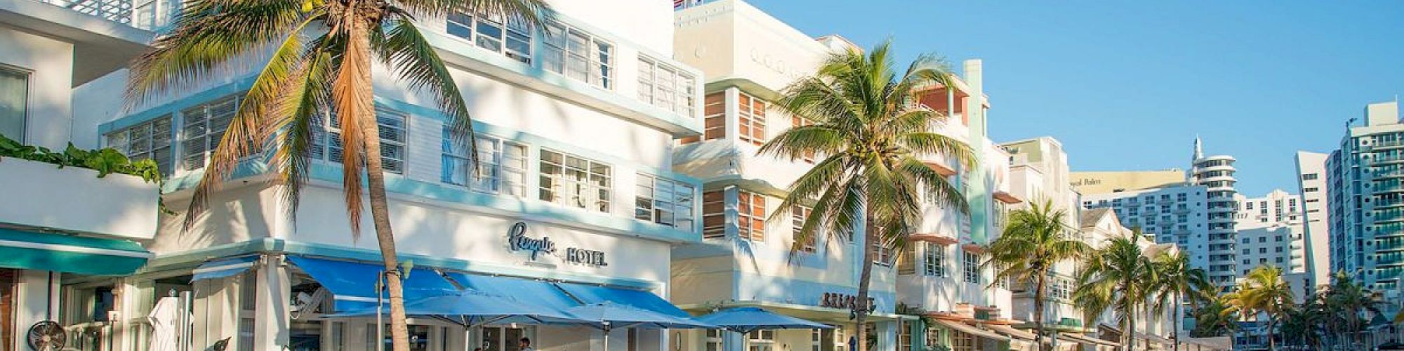 A sunny street scene with palm trees, Art Deco buildings, outdoor dining, and parked cars in a coastal city.