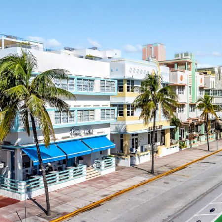 A sunny street with pastel buildings, palm trees, and a blue awning storefront along a bike-friendly, shaded seaside road.