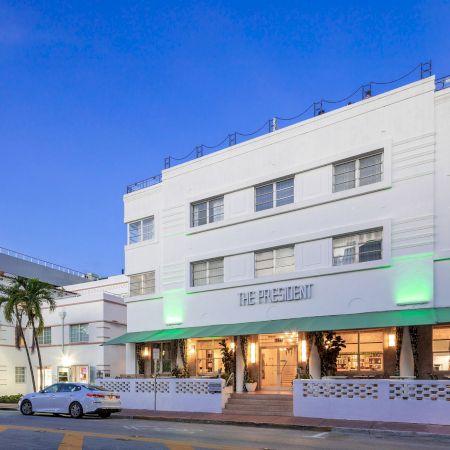 A white, three-story building with a mint green awning reads &ldquo;The Crescent,&rdquo; parked cars line the street, palm trees, and a clear blue sky.