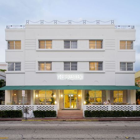 A white multi-story Art Deco-style hotel with a green awning, large windows, and a small front entrance; plants line the building.