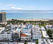 Aerial view of a beachfront hotel complex with palm trees, a sandy shore, calm blue ocean, and small boats on the horizon.