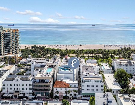 Aerial view of a beachfront hotel complex with palm trees, a sandy shore, calm blue ocean, and small boats on the horizon.