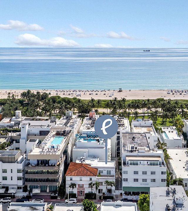 Aerial view of a beachfront hotel complex with palm trees, a sandy shore, calm blue ocean, and small boats on the horizon.