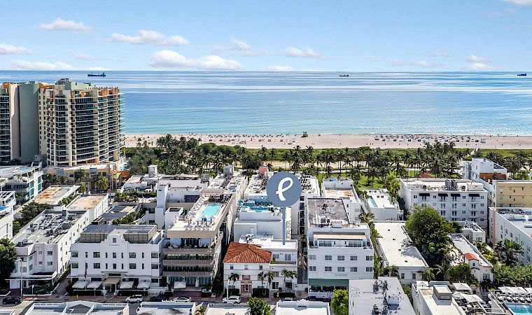 Aerial view of a beachfront hotel complex with palm trees, a sandy shore, calm blue ocean, and small boats on the horizon.