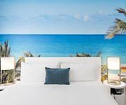 White outdoor daybed lounge with pillows, palm tree, and a wide blue ocean view under a clear sky.