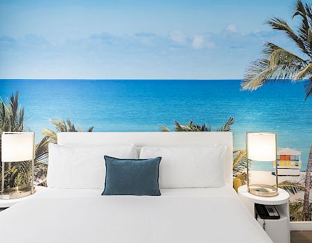 White outdoor daybed lounge with pillows, palm tree, and a wide blue ocean view under a clear sky.