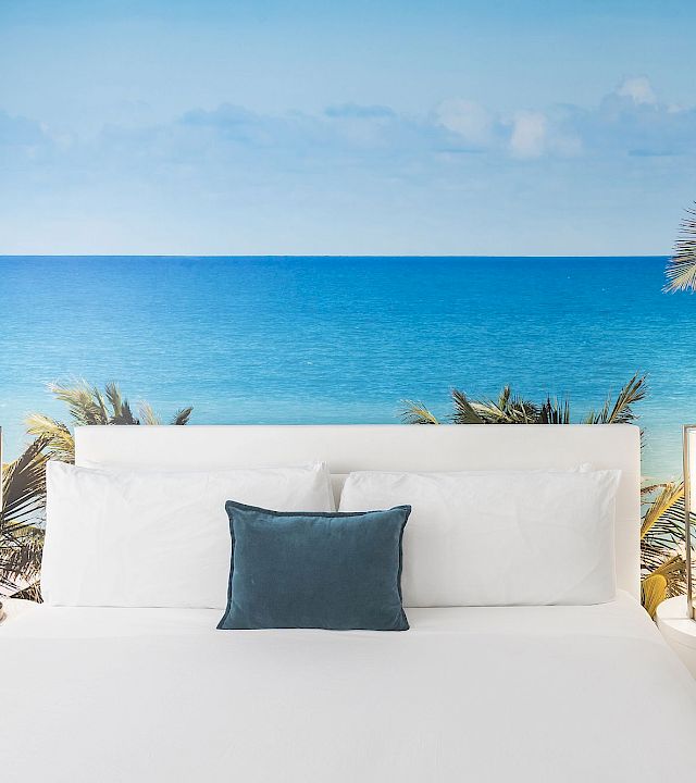 White outdoor daybed lounge with pillows, palm tree, and a wide blue ocean view under a clear sky.