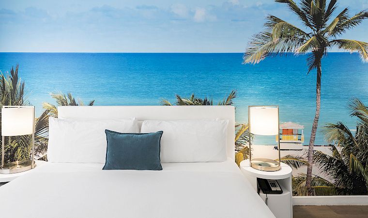 White outdoor daybed lounge with pillows, palm tree, and a wide blue ocean view under a clear sky.