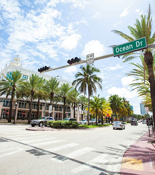 A sunny street corner with the “Ocean Dr” sign, palm trees, cars, and traffic lights under a blue sky.