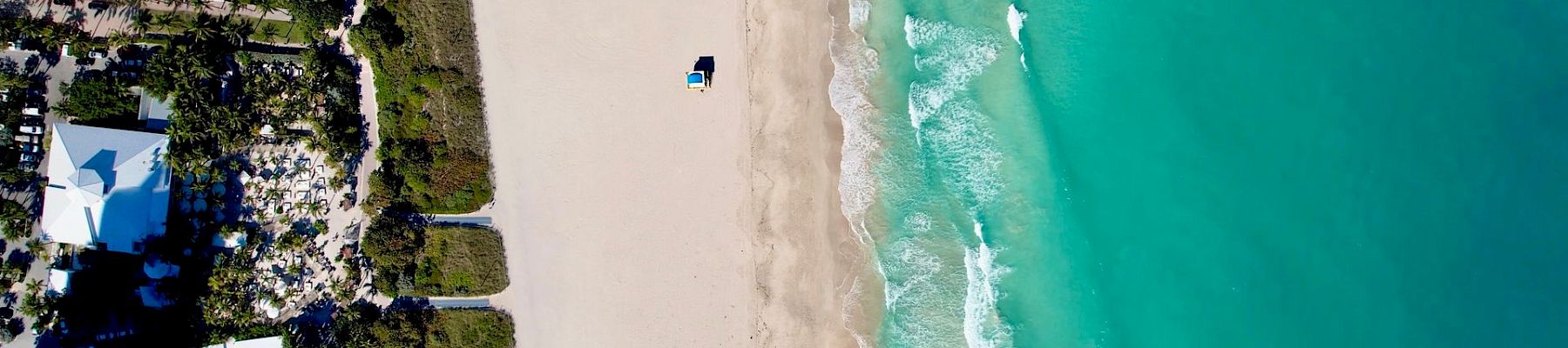 Aerial view of a beachfront resort beside a long sandy shore, with turquoise ocean waves rolling in.