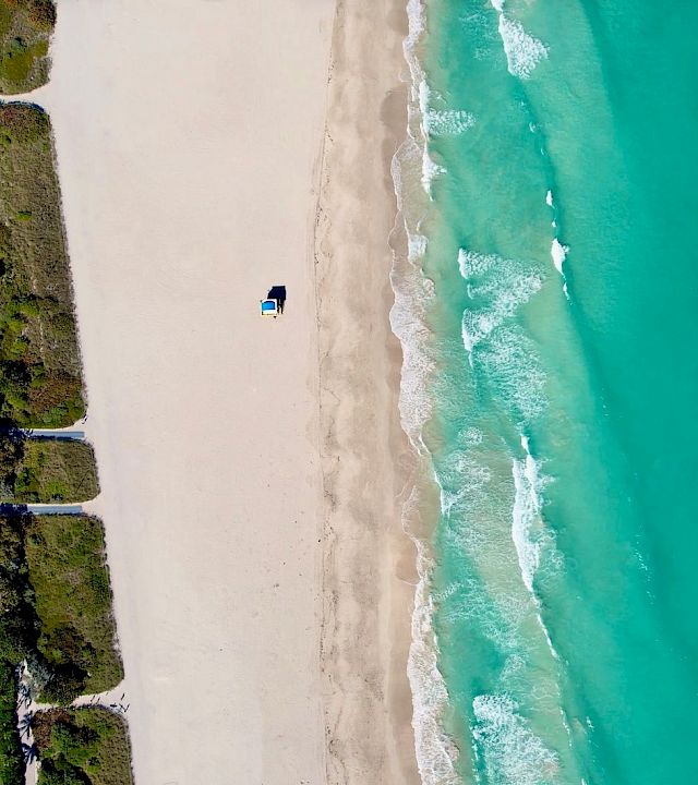 Aerial view of a beachfront resort beside a long sandy shore, with turquoise ocean waves rolling in.
