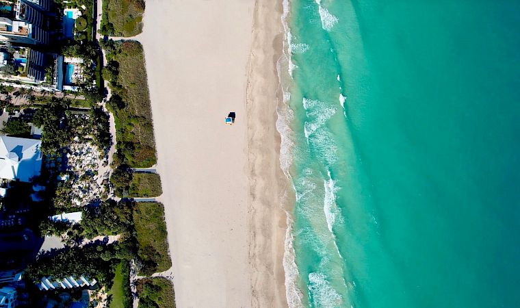 Aerial view of a beachfront resort beside a long sandy shore, with turquoise ocean waves rolling in.
