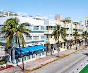 A sunny city street lined with palm trees, a modern hotel building, and distant towers under a blue sky.