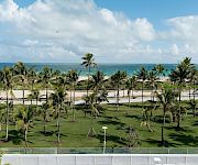 Tropical seaside view: palm-lined garden overlooking a sandy beach and turquoise ocean under big fluffy clouds.