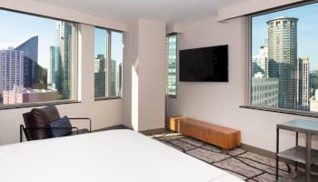 The image shows a modern hotel room with large windows offering a city skyline view, a bed, a TV, a chair, and a small table.