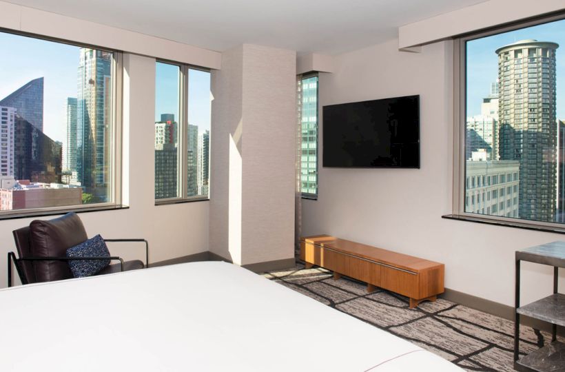 The image shows a modern hotel room with large windows offering a city skyline view, a bed, a TV, a chair, and a small table.