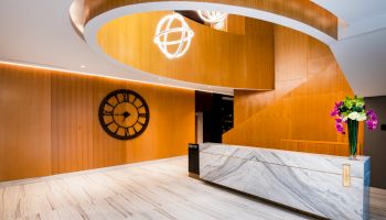 A modern lobby with a curved wooden staircase, a large clock on the wall, a marble reception desk, and a vase with flowers on it.