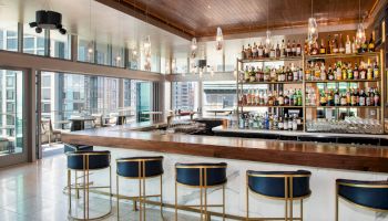 A modern bar with a wooden counter, lined with high-back stools, a well-stocked liquor shelf, and large windows offering city views.