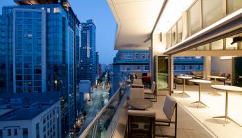 A modern cityscape at dusk, with a stylish outdoor balcony seating area overlooking tall buildings and a brightly lit street below.