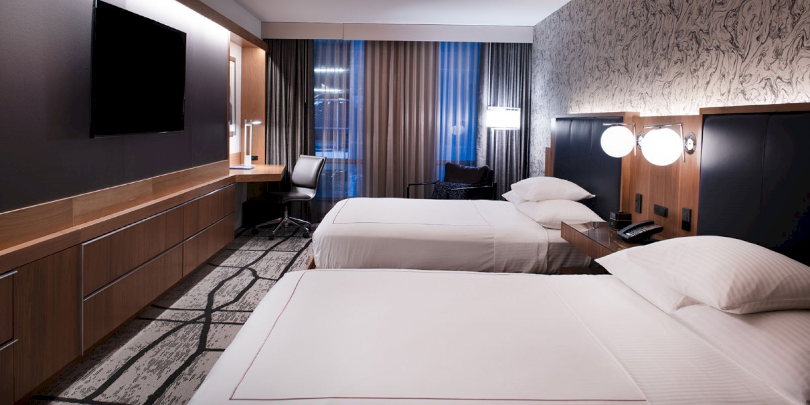 A modern hotel room with two neatly made beds, a wall-mounted TV, desk, and large window with drapes. The room has stylish decor and lighting fixtures.