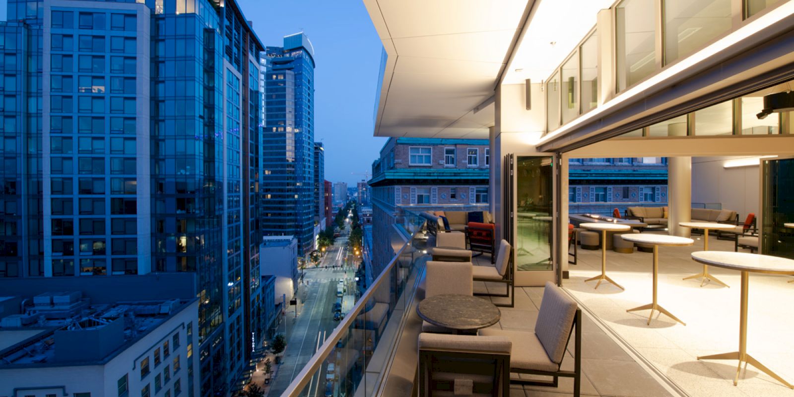 This image shows an upscale outdoor balcony with seating and tables, overlooking a modern city street with tall buildings during twilight.