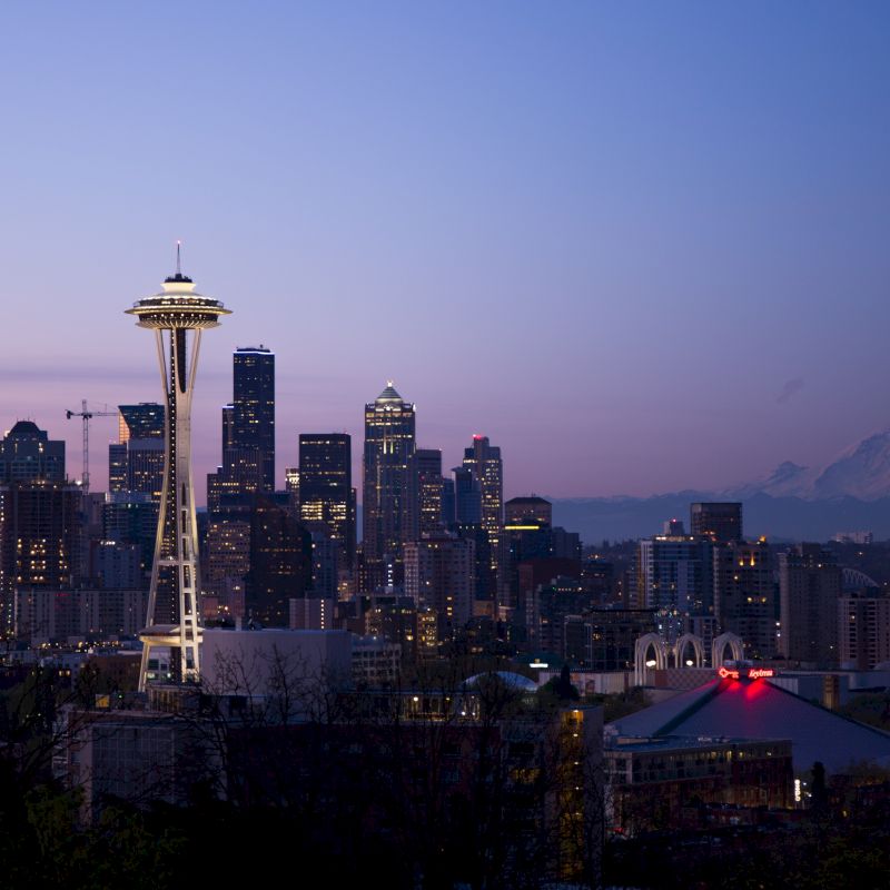 A cityscape at dusk featuring a prominent tower, a skyline of buildings, and a distant mountain under a clear sky.
