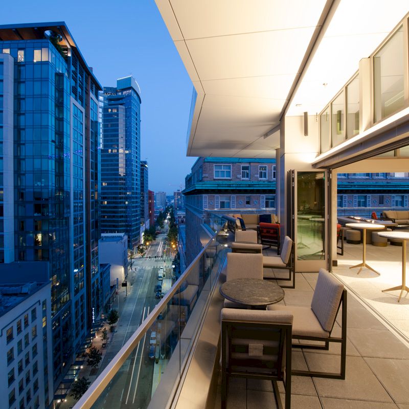 The image shows a modern balcony overlooking a cityscape with tall buildings, featuring seating and tables, and large glass windows.