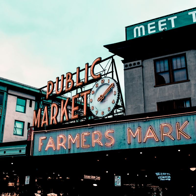 The image shows a neon sign for "Public Market" and "Farmers Market" with a clock, in front of buildings that appear to house a hotel and various shops.