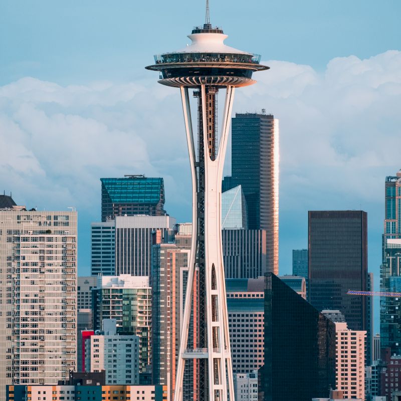 The image shows the Space Needle tower in Seattle, with the city's downtown skyscrapers and buildings in the background, against a cloudy sky.