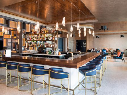 The image shows a modern bar with a stocked liquor shelf, stylish blue and gold bar stools, pendant lights, and a lounge area in the background.