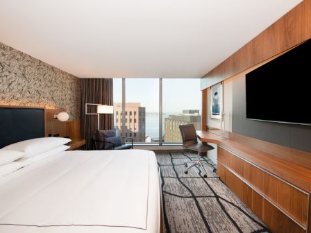 A modern hotel room with a large bed, wall-mounted TV, work desk, and floor-to-ceiling windows offering a city view.