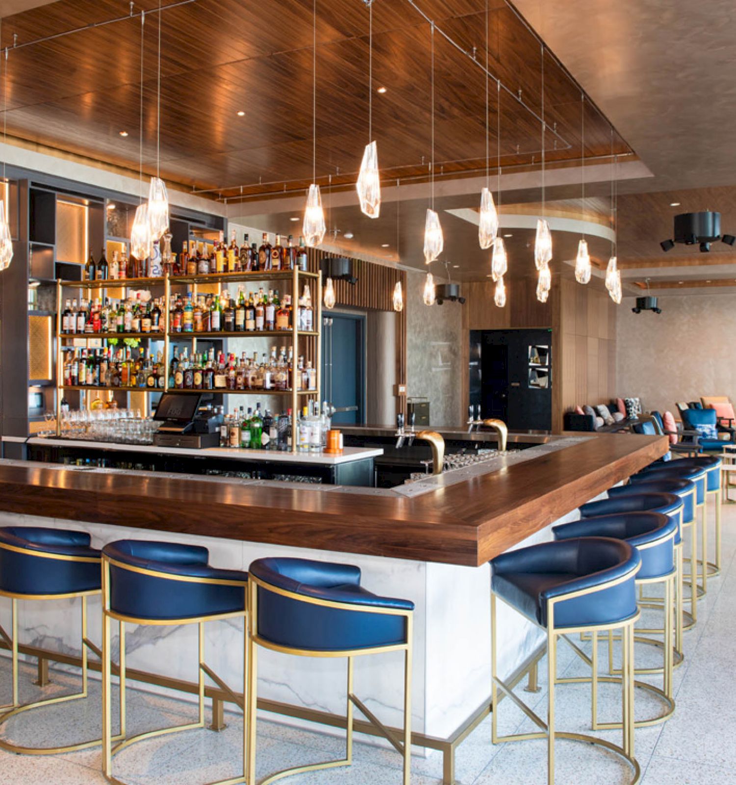 This image shows a modern bar with a well-stocked liquor display, blue bar stools, hanging lights, and a lounge area with sofas and tables.