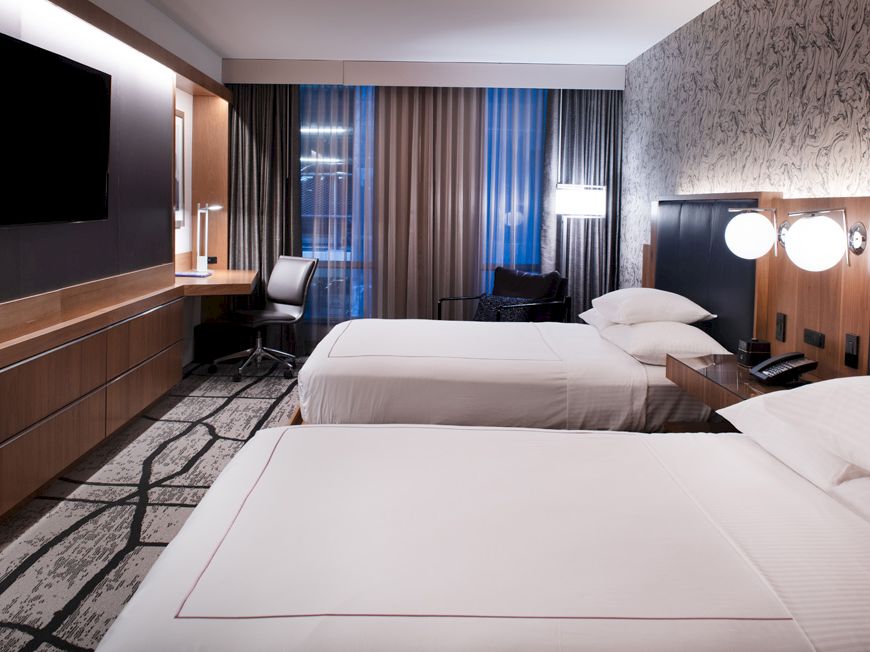 A modern hotel room with two double beds, wall-mounted TV, desk, chair, large window with curtains, and ambient lighting on a textured wall.