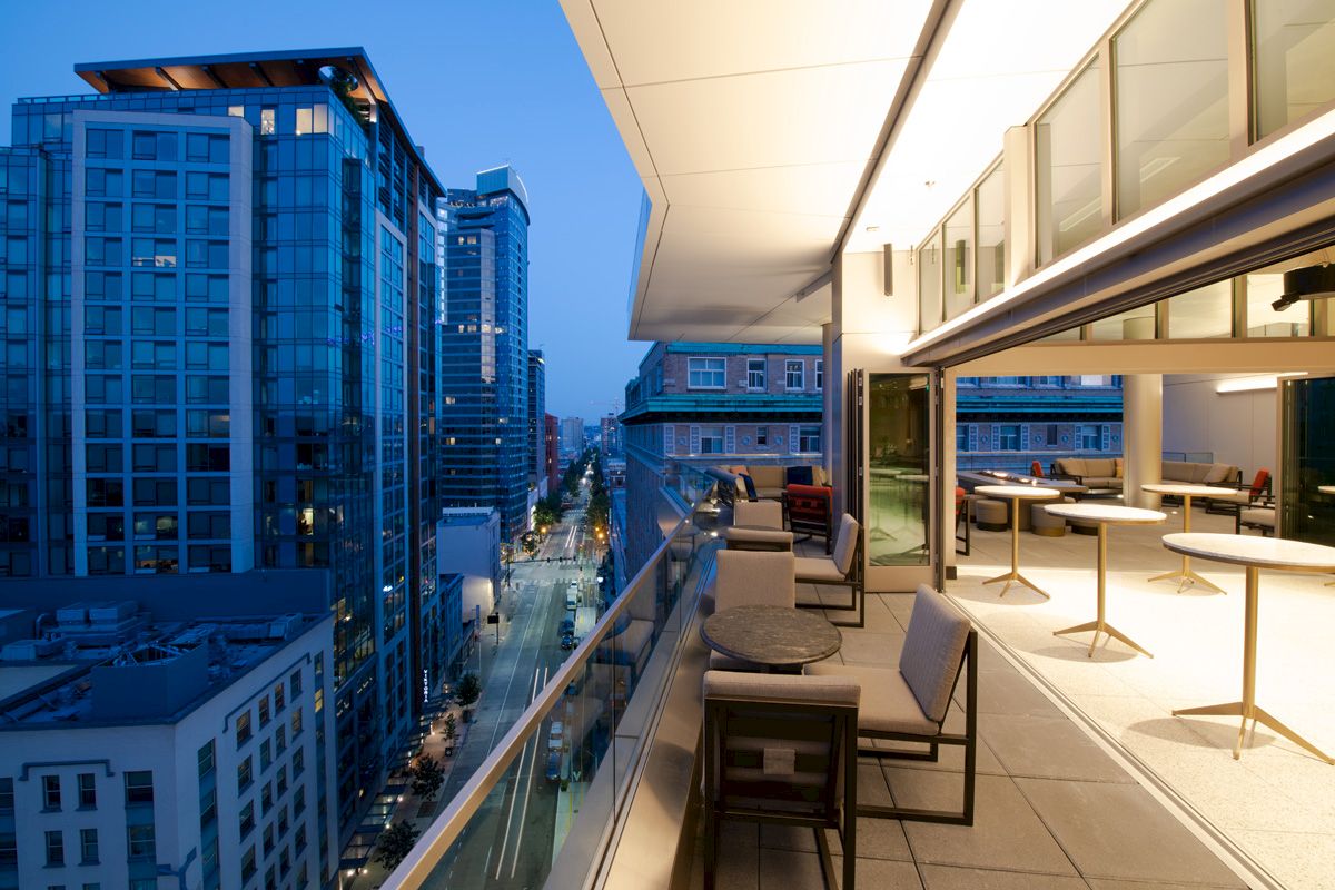 A modern rooftop patio at dusk, featuring tables, chairs, and a view of an urban street lined with high-rise buildings.
