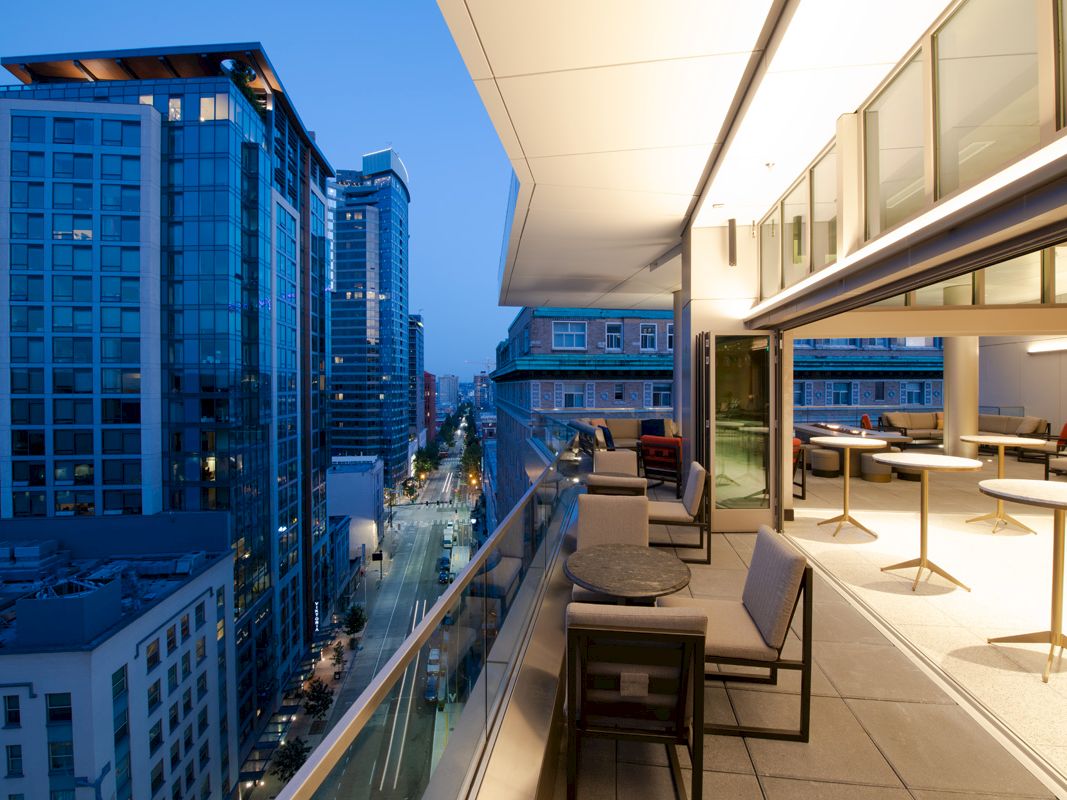 A modern rooftop patio at dusk, featuring tables, chairs, and a view of an urban street lined with high-rise buildings.