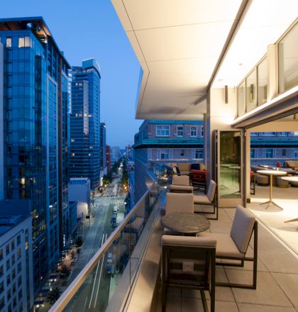 A modern urban balcony with seating overlooks a city street lined with tall buildings at dusk, with indoor seating visible through glass doors.