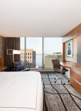 A modern hotel room with a bed, desk, chair, wall-mounted TV, and a window offering a city view is seen in the image.
