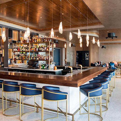 The image shows a modern bar with a wooden counter, blue and gold bar stools, pendant lights, and a fully stocked back bar, all in a stylish lounge.