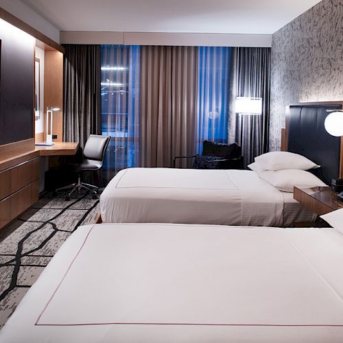 A modern hotel room with two double beds, a flat-screen TV, a desk with a chair, and stylish décor including wall art and lamps.