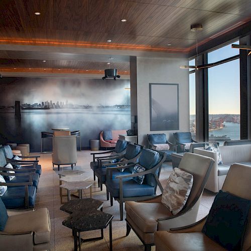 A modern lounge area with comfortable seating, large windows offering a scenic view of water and hills, and artwork on the wall.