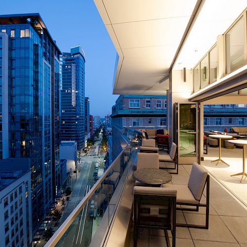 An upscale balcony overlooking a modern cityscape with high-rise buildings during dusk, featuring outdoor seating and lighting.