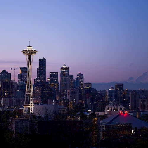 A cityscape at dusk with a prominent tower and mountain in the background, surrounded by a mix of modern buildings and a glowing sky.