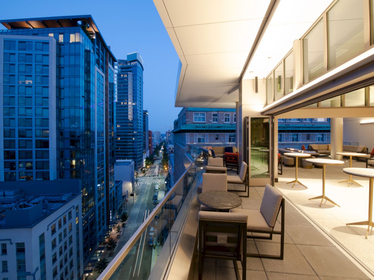 A cityscape during dusk featuring high-rise buildings and a modern balcony furnished with tables and chairs overlooking the urban view.