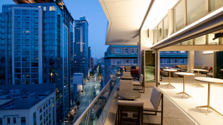 A cityscape during dusk featuring high-rise buildings and a modern balcony furnished with tables and chairs overlooking the urban view.