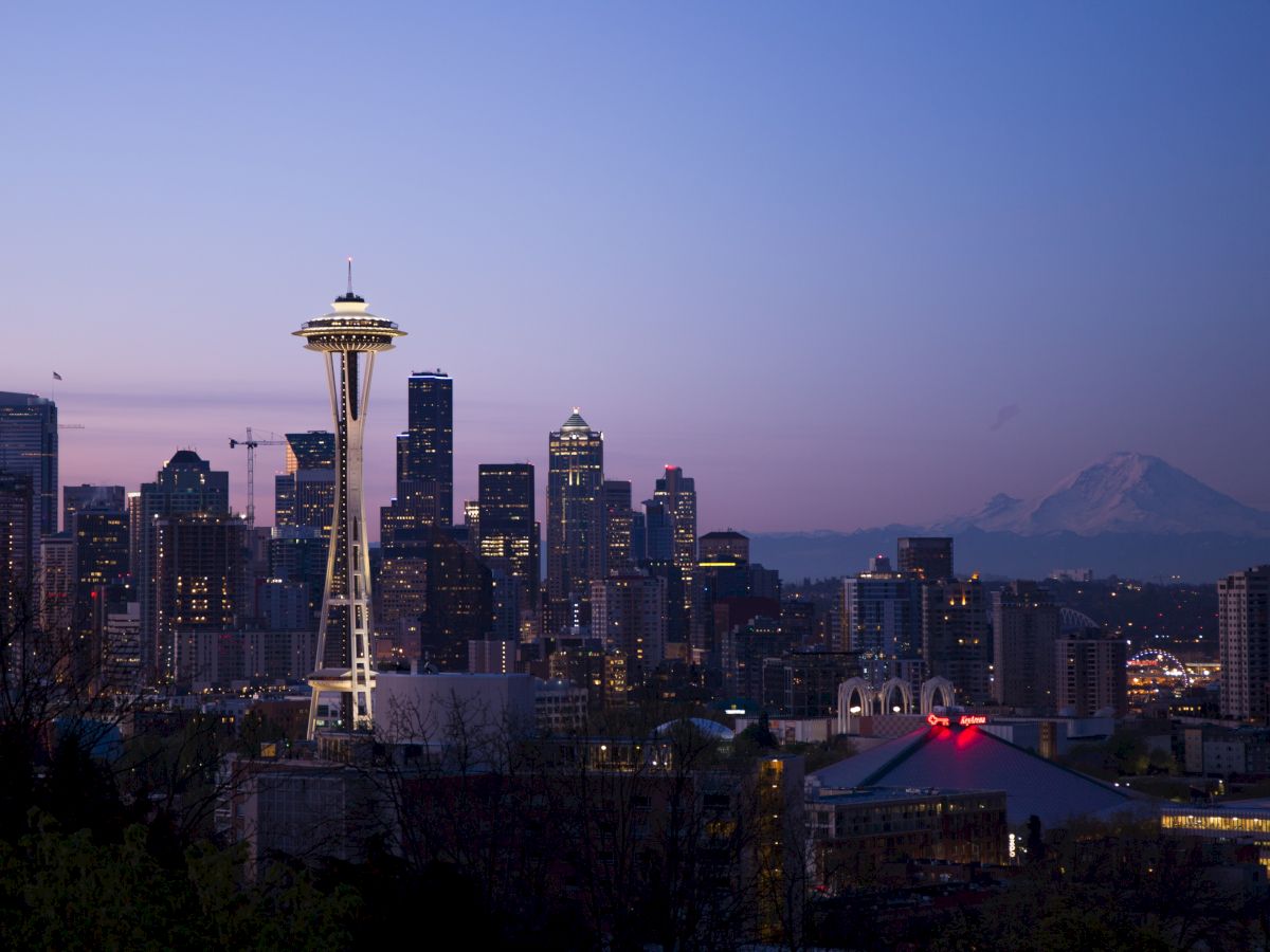 A city skyline at dusk with tall buildings, the Space Needle, and a distant mountain under a clear sky.