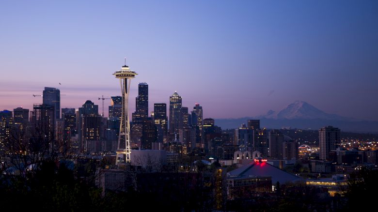A city skyline at dusk with tall buildings, the Space Needle, and a distant mountain under a clear sky.