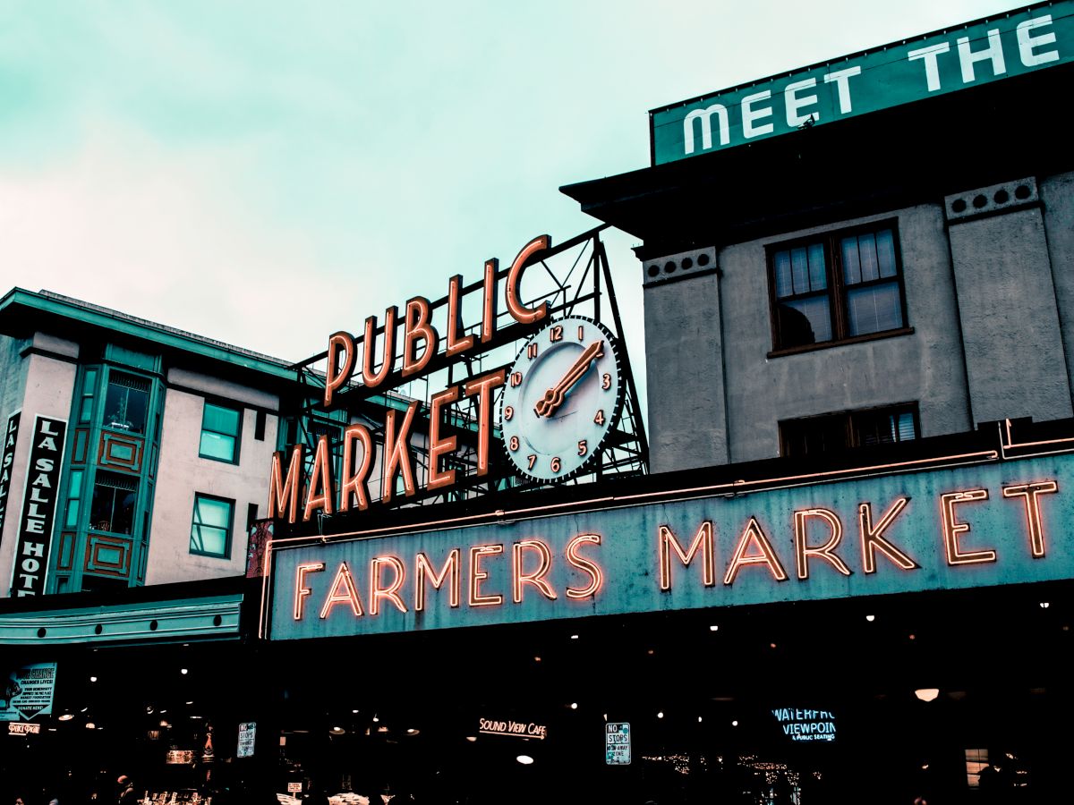 The image shows a neon sign for a public market and farmers market, with an iconic clock, located on buildings with visible windows and signs.
