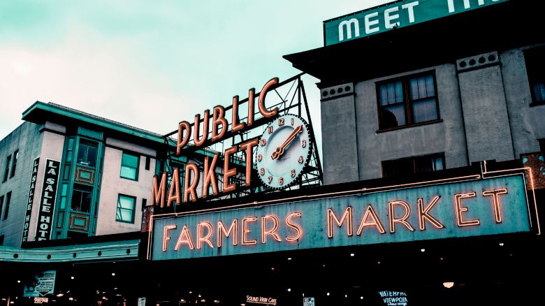 The image shows a neon sign for a public market and farmers market, with an iconic clock, located on buildings with visible windows and signs.
