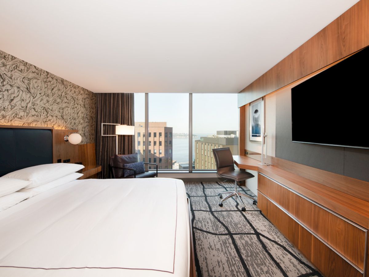 Modern hotel room with a large bed, wall-mounted TV, desk, chair, floor-to-ceiling window, and city view.